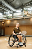Vertical full length portrait of young woman in wheelchair holding racket during practice at indoor sports court