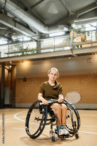 Vertical full length portrait of young woman in wheelchair holding racket during practice at indoor sports court