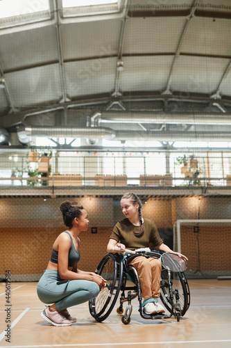 Vertical full length portrait of young woman in wheelchair talking to coach or teammate during sports practice at indoor court
