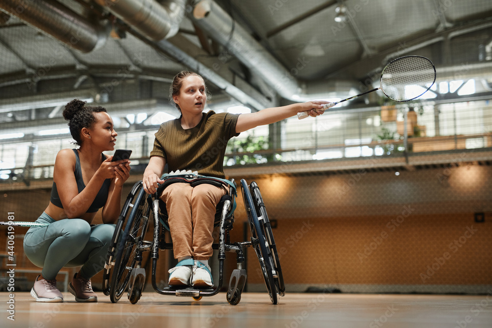 Fototapeta premium Full length portrait young woman in wheelchair talking to coach during badminton practice at sports court, copy space