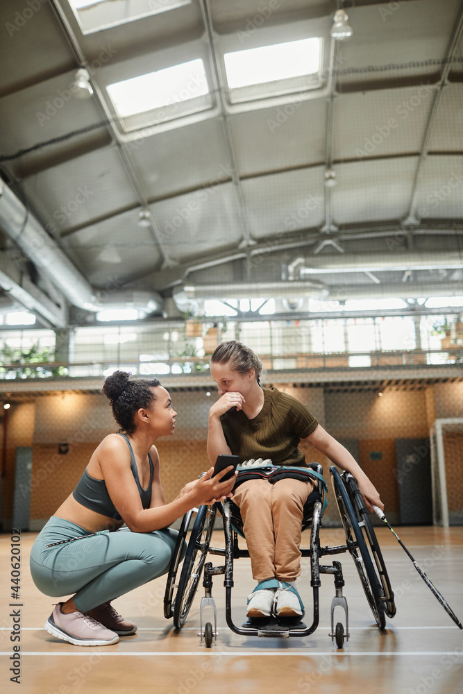 Vertical full length portrait young woman in wheelchair talking to friend during badminton practice at sports court Vertical full length portrait young woman in wheelchair talking to friend during badminton practice at sports court
