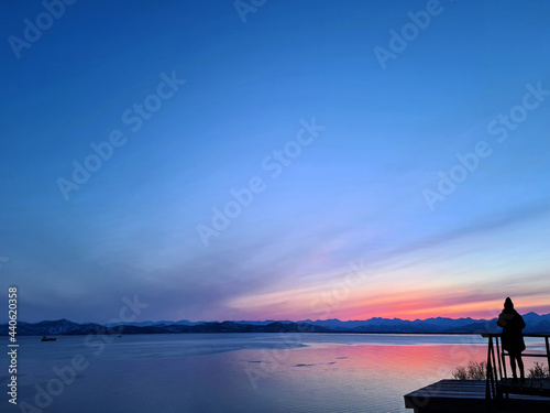 Sunset view of the Avacha Bay with a silhouette of a girl standing on a balcony. Ships standing in the roadstead, mountains in the background. Petropavlovsk-Kamchatsky, Kamchatka Peninsula, Russia photo