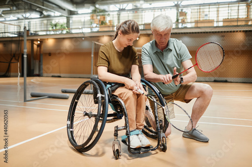 Full length portrait of smiling young woman in wheelchair talking to coach during badminton practice at sports court