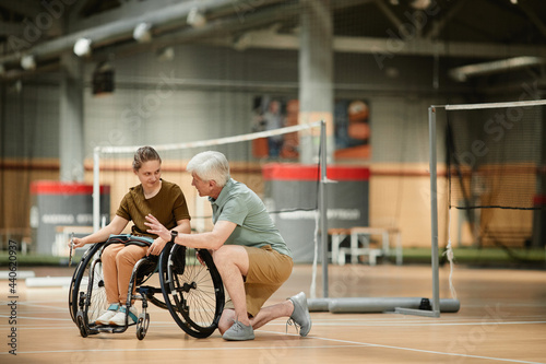 Full length portrait of senior coach talking to young woman in wheelchair during badminton practice in sports court, copy space