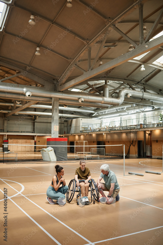 Vertical wide angle portrait of mature coach talking to young woman in wheelchair during badminton practice in sports court, copy space