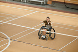 Wide angle view at young woman in wheelchair playing badminton during sports practice at indoor court, copy space