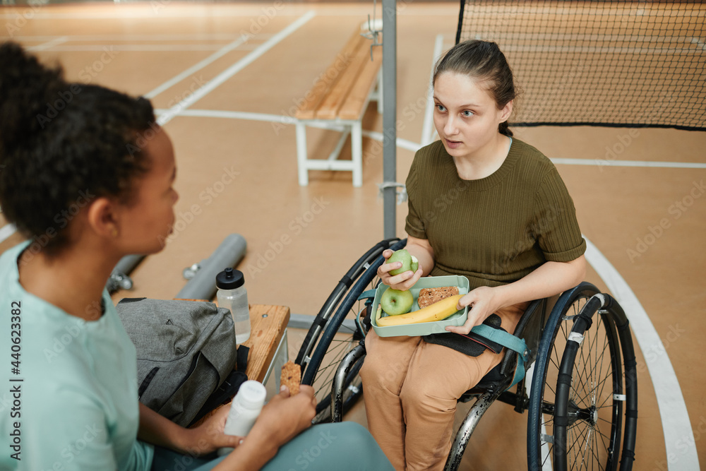 Obraz premium High angle portrait of young woman in wheelchair eating healthy snack with friend at indoor sports court, copy space