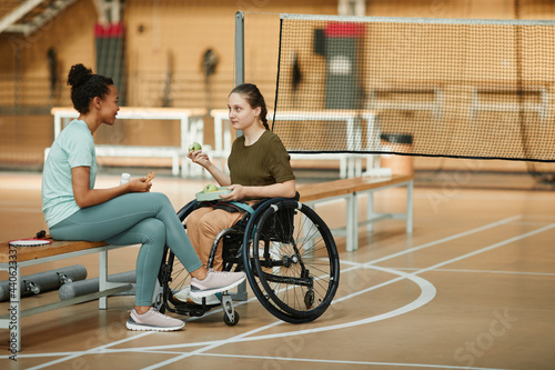 Full length portrait of young woman in wheelchair sharing healthy snack with friend at indoor sports court, copy space