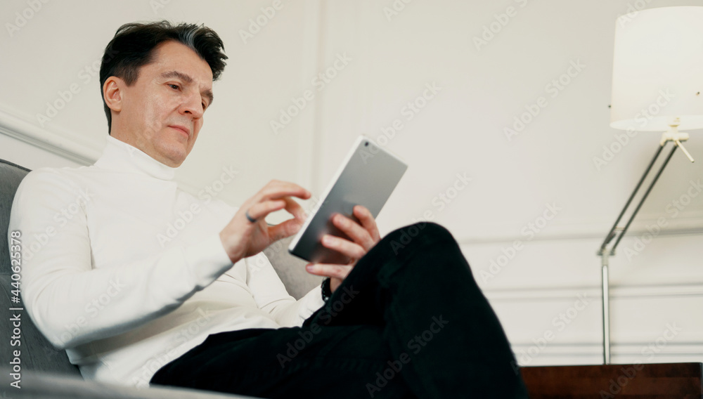 An adult gray-haired man of European appearance types a message to a client on a tablet. The male manager works in the gadget. Online distance learning.