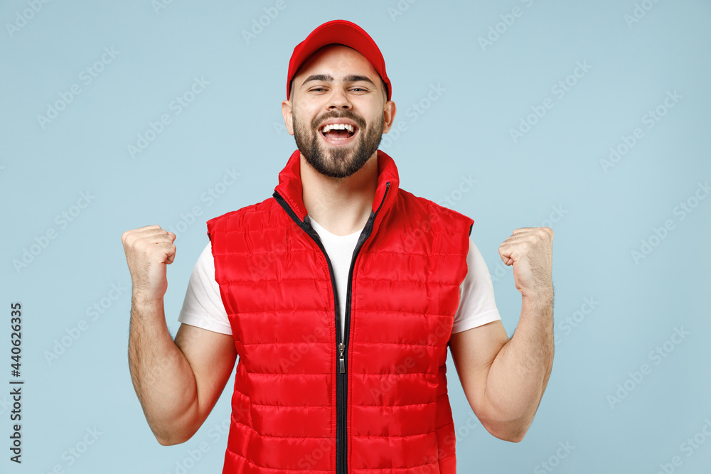 Naklejka premium Professional delivery guy employee man in red cap white T-shirt vest uniform workwear work as dealer courier winner celebrate isolated on pastel blue color background studio portrait. Service concept.