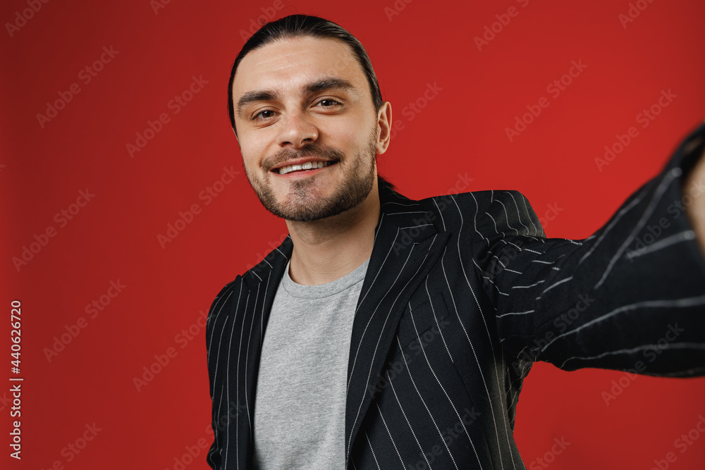 Close up young smiling happy businessman latin man in black striped jacket grey shirt doing selfie shot on mobile phone isolated on red background studio studio portrait. Concept of people lifestyle. Close up young smiling happy businessman latin man in black striped jacket grey shirt doing selfie shot on mobile phone isolated on red background studio studio portrait. Concept of people lifestyle.