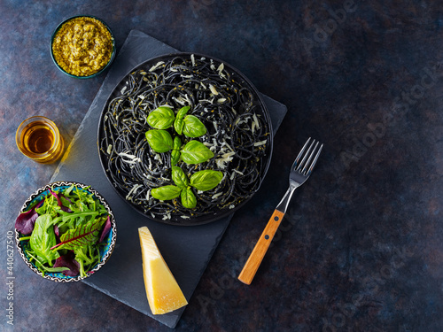 Black pasta with parmesan cheese, basil and pesto sauce. Black spaghetti with cuttlefish ink on a dark background. Pasta with grated cheese and fresh greens mix salad. Top view. Copy space