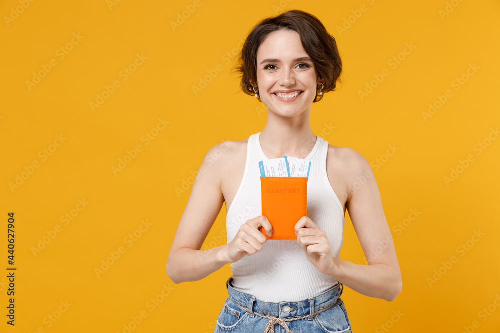 Traveler smiling happy tourist woman 20s wearing white tank top shirt hold passport tickets isolated on yellow color background. Passenger travel abroad on weekends getaway Air flight journey concept. Traveler smiling happy tourist woman 20s wearing white tank top shirt hold passport tickets isolated on yellow color background. Passenger travel abroad on weekends getaway Air flight journey concept.