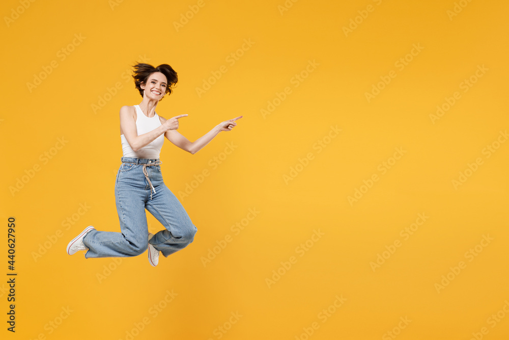 Full length young fun happy amazed woman 20s with bob haircut wearing white tank top shirt jumping high pointing index finger aside on copy space area mock up workspace isolated on yellow background.