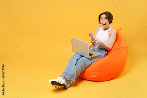 Young surprised woman 20s with bob haircut in white tank top shirt point index finger on laptop pc computer chat online browsing surfing internet sit in orange bag chair isolated on yellow background photo