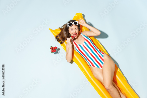 Top view sexy woman slim body wear striped one-piece swimsuit lies on inflatable mattress hotel pool eats red berries isolated on pastel blue color background Summer vacation sea rest sun tan concept