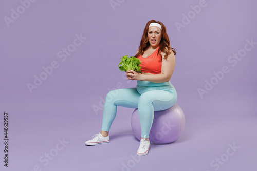 Full length indignant sad young chubby overweight plus size big fat woman in red top warm up train sit on fit ball hold lettuce leaves isolated on purple color background gym Workout sport concept
