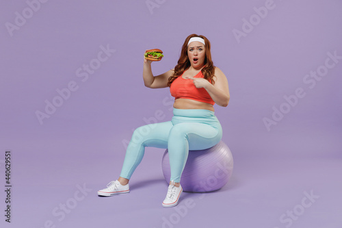 Full length indignant young chubby overweight plus size big fat woman in red top warm up train sit on fit ball point finger on fast food burger isolated on purple background gym Workout sport concept