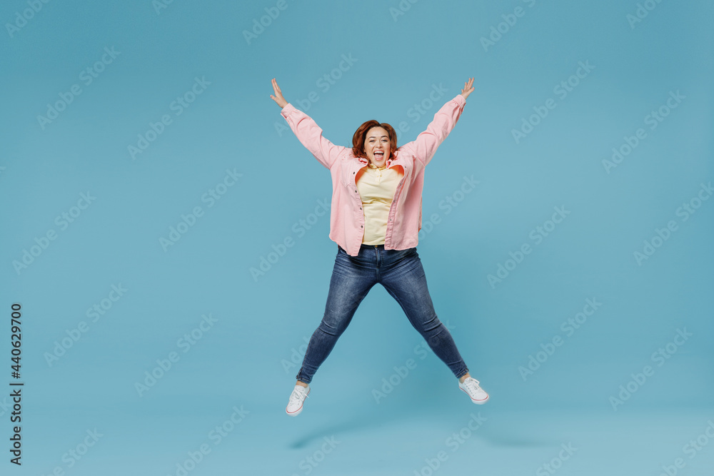 Full length young joyful smiling redhead chubby overweight woman 30s wearing in pink shirt jeans jumping high with oustretched hands looking camera have fun isolated on pastel blue background studio.