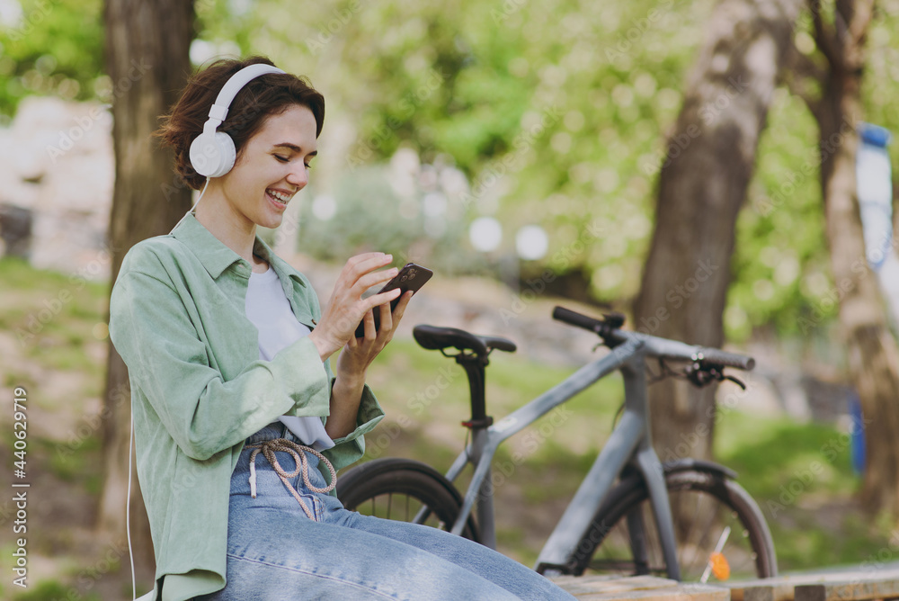 Young woman in casual green jacket jeans headphones sit on bench near bicycle bike in city spring park outdoors hold mobile cell phone listen to music chat online People active urban youthful concept.