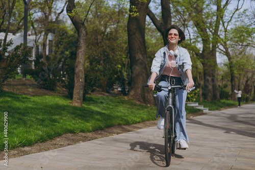 Full length young sporty woman 20s wear casual jeans clothes headphones riding bicycle bike on sidewalk in city spring park outdoors, look aside. People active urban healthy lifestyle cycling concept