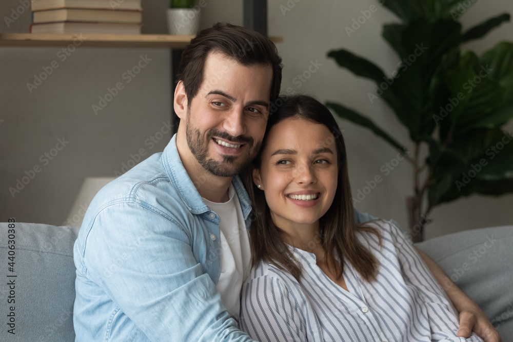 Portrait of sincere happy dreamy young family couple cuddling, resting on comfortable sofa. Affectionate loving millennial man and woman looking in distance, imagining planning future at home.