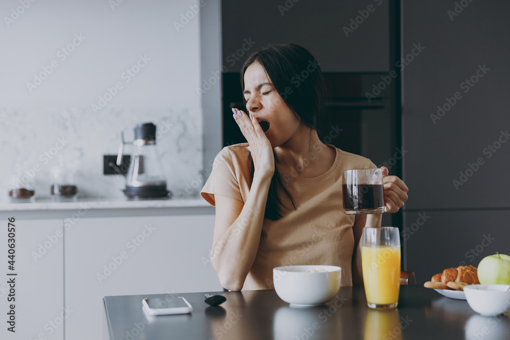 Young tired sleepy housewife woman 20s in casual beige t-shirt eat breakfast hold cup of tea drink coffee yawning cover mouth cook food in light kitchen at home alone Healthy diet lifestyle concept. Young tired sleepy housewife woman 20s in casual beige t-shirt eat breakfast hold cup of tea drink coffee yawning cover mouth cook food in light kitchen at home alone Healthy diet lifestyle concept.