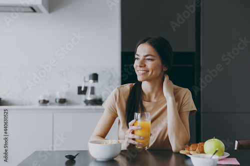 Young dreamful brunette housewife happy woman 20s wearing casual clothes beige shirt eat oatmeal porridge muesli drink juice cooking food in light kitchen at home alone Healthy diet lifestyle concept