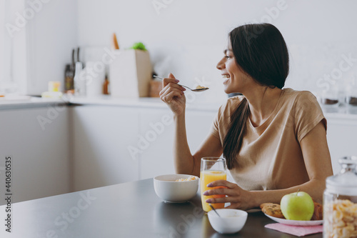Side view young happy smiling housewife woman 20s wearing casual clothes beige shirt eat oatmeal porridge muesli in morning cooking food in light kitchen at home alone. Healthy diet lifestyle concept