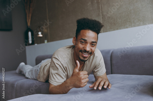 Full length fun young african american man in casual beige t-shirt sweatpants lay down on grey sofa show thumb up indoors partment look camera resting on weekends staying at home during quarantine photo