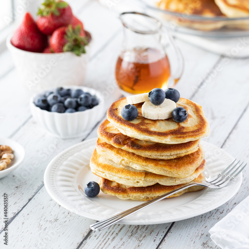 A stack of buttermilk pancakes topped with blueberries and banana, ready for syrup to be poured on top.