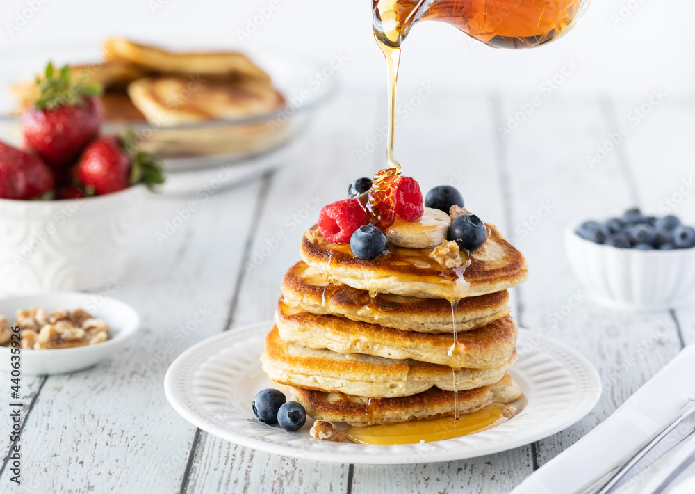 A stack of buttermilk pancakes served with berries and bananas with syrup being poured on top. A stack of buttermilk pancakes served with berries and bananas with syrup being poured on top.