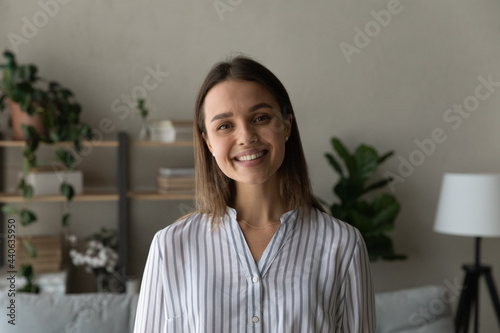 Portrait of sincere beautiful young female homeowner with perfect white smile posing in modern living room. Smiling joyful candid millennial generation woman looking at camera, holding video call.