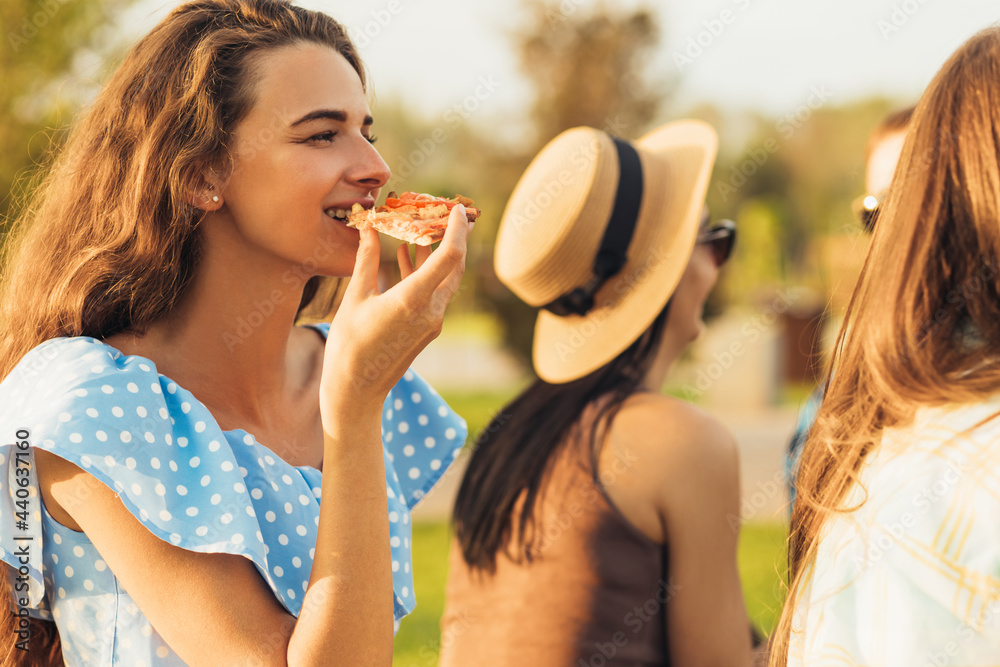 Three smiling beautiful girlfriends, spend time together, sit on the lawn, eat pizza and drink drinks on a picnic
