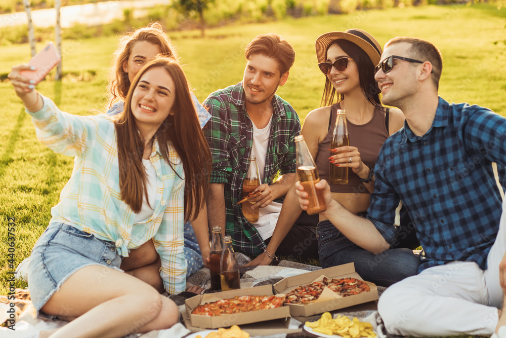 Group of young people taking selfie while eating pizza, friends are sitting around during an outdoor party on the grass