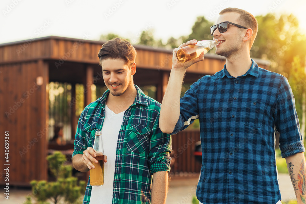 Fototapeta premium Two young male friends walking in the park in summer on a sunny day, talking and drinking beer drinks