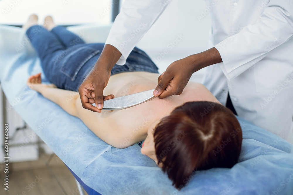 Massage with special metal scraper device. Rehabilitation concept. Cropped shot of hands of African male physiotherapist conducting back massage for young lady using gouache scraper.