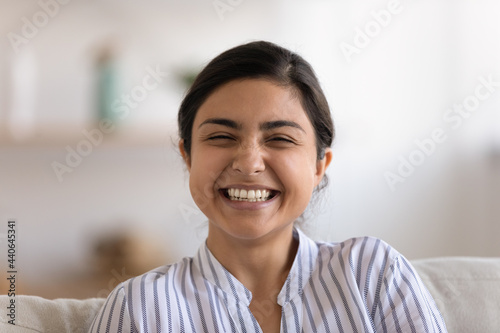 Close up profile picture of overjoyed millennial teen Indian female look at camera relaxing in cozy home. Headshot portrait of smiling young mixed race ethnicity woman have fun rest in living room.