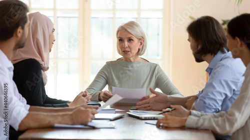 Middle aged businesswoman team leader holding corporate meeting with diverse employees, mature coach mentor holding documents, training or teaching staff, discussing project statistics in boardroom