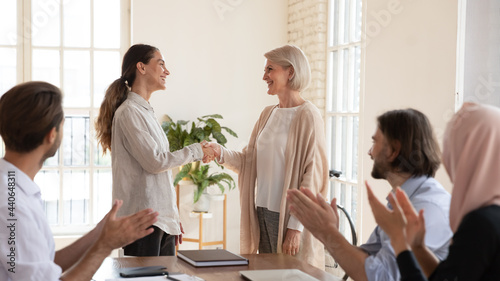 Mature executive shaking successful businesswoman hand at corporate meeting, middle aged team leader greeting new employee, young intern or thanking for good work result, colleagues applauding