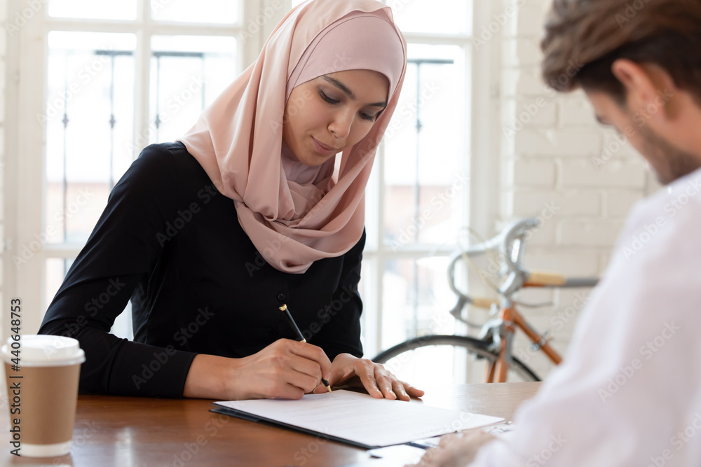 Fototapeta premium Asian muslim businesswoman wearing hijab signing job contract or agreement with business partner at meeting, saleswoman putting signature on legal documents, making investment or insurance deal