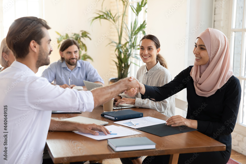 Smiling Asian muslim businesswoman shaking hand of businessman making agreement at meeting in boardroom, group negotiation, diverse business partners handshaking, celebrating contract signing Smiling Asian muslim businesswoman shaking hand of businessman making agreement at meeting in boardroom, group negotiation, diverse business partners handshaking, celebrating contract signing