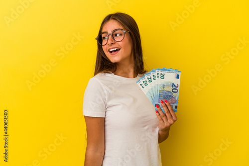 Young caucasian woman holding bills isolated on yellow background looks aside smiling, cheerful and pleasant.