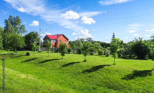 A lawn with green grass and small trees in front of a two-story red brick house. Cottage on a sunny summer day.