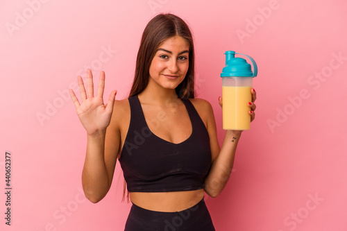 Young caucasian woman holding a protein shake isolated on yellow background smiling cheerful showing number five with fingers.