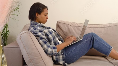 Beautiful dark-skinned woman in casual clothes working at home online lying on the couch. A large palm tree plant in a flower pot.