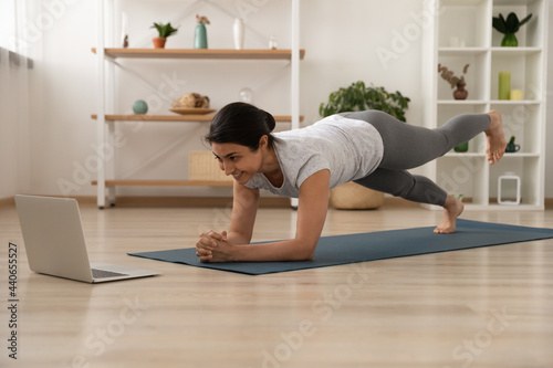 Happy millennial Indian female in sportswear on mat at home practice yoga stretching with online lesson or class on computer. Smiling ethnic woman train do workout watch video session on laptop.