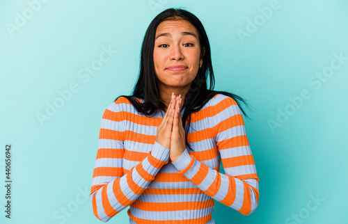 Young Venezuelan woman isolated on blue background holding hands in pray near mouth, feels confident.