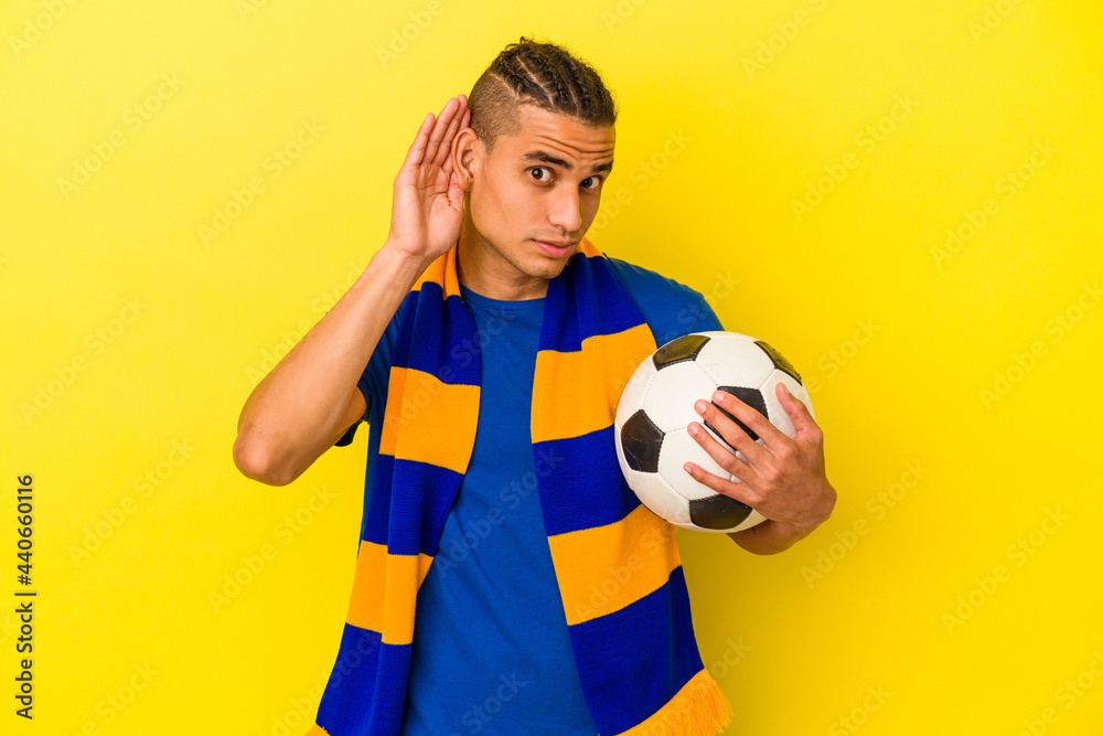 Young venezuelan man watching soccer isolated on yellow background trying to listening a gossip.