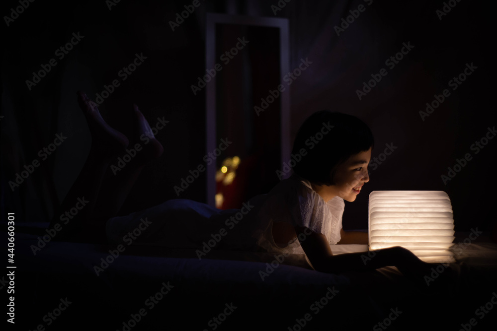 Bella niña asiática leyendo libro luminoso en la cama Bella niña asiática leyendo libro luminoso en la cama